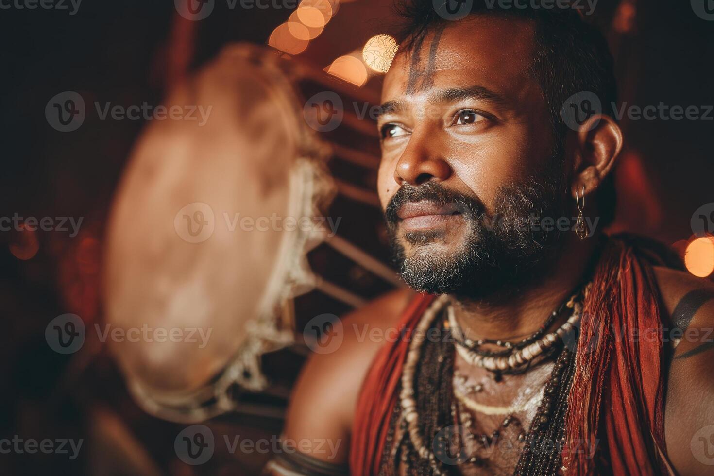 Bearded man with a thoughtful expression, adorned in traditional accessories, gazes to the side. Dim lighting highlights beads and a large drum in the background photo