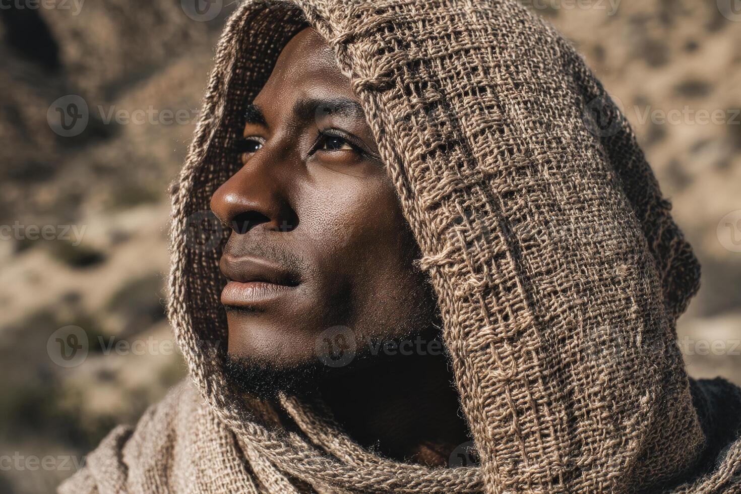 A contemplative person gazes upward, wrapped in a textured, brown hooded shawl. The sunlight casts gentle shadows on their face, highlighting the serene expression against a blurred natural backdrop photo