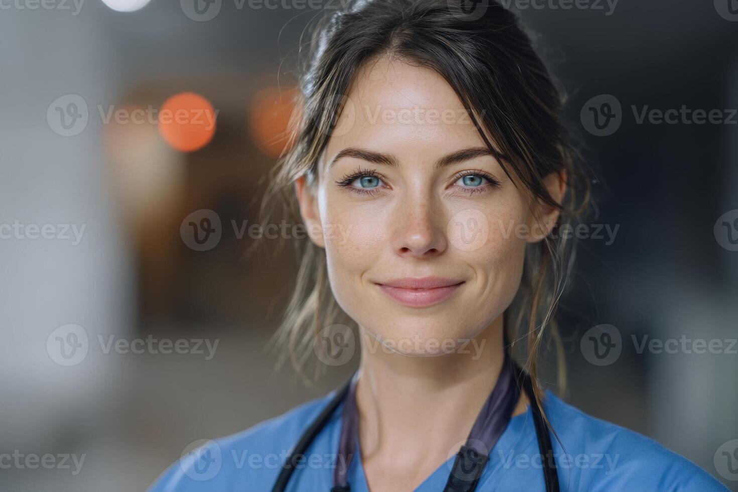Smiling woman wearing light blue medical attire with a stethoscope around her neck, dark brown hair tied back, standing in a softly focused indoor setting with warm blurred lights in the distance photo