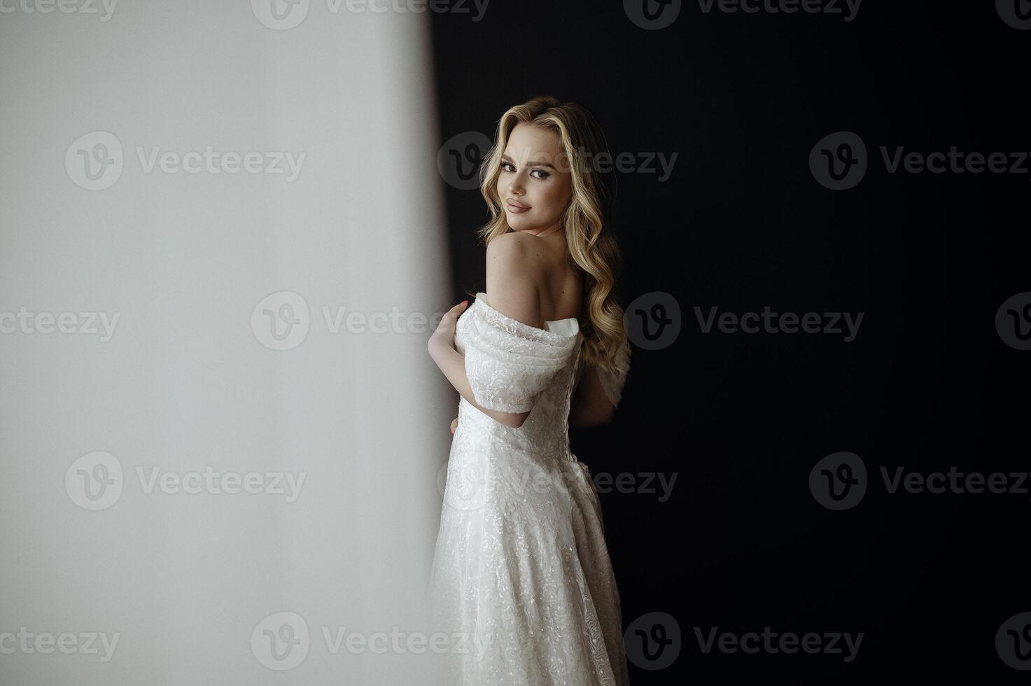 Elegant bridal gown showcased in a minimalist setting with a model in a chic pose during an indoor shoot. image of a blonde bride against a dark interior. smiles happily and looks at the camera photo