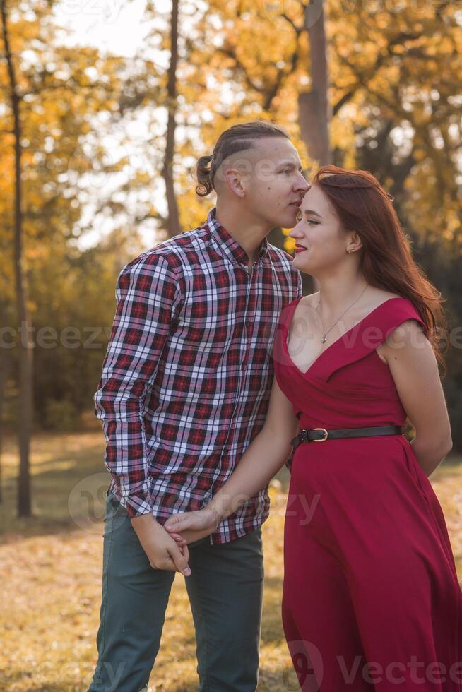 Portrait of a young couple in love kissing and hugging in an autumn park. Girl in a red dress against the backdrop of golden autumn. Concept of romance, falling in love photo