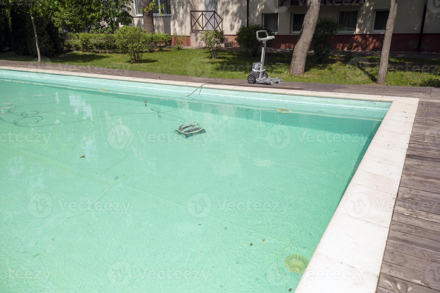 Swimming pool with wooden decking in the hotel garden, and water cleaning equipment. The pool has a pool cleaning machine. The equipment is in the water and nearby. The hotel is in the background photo