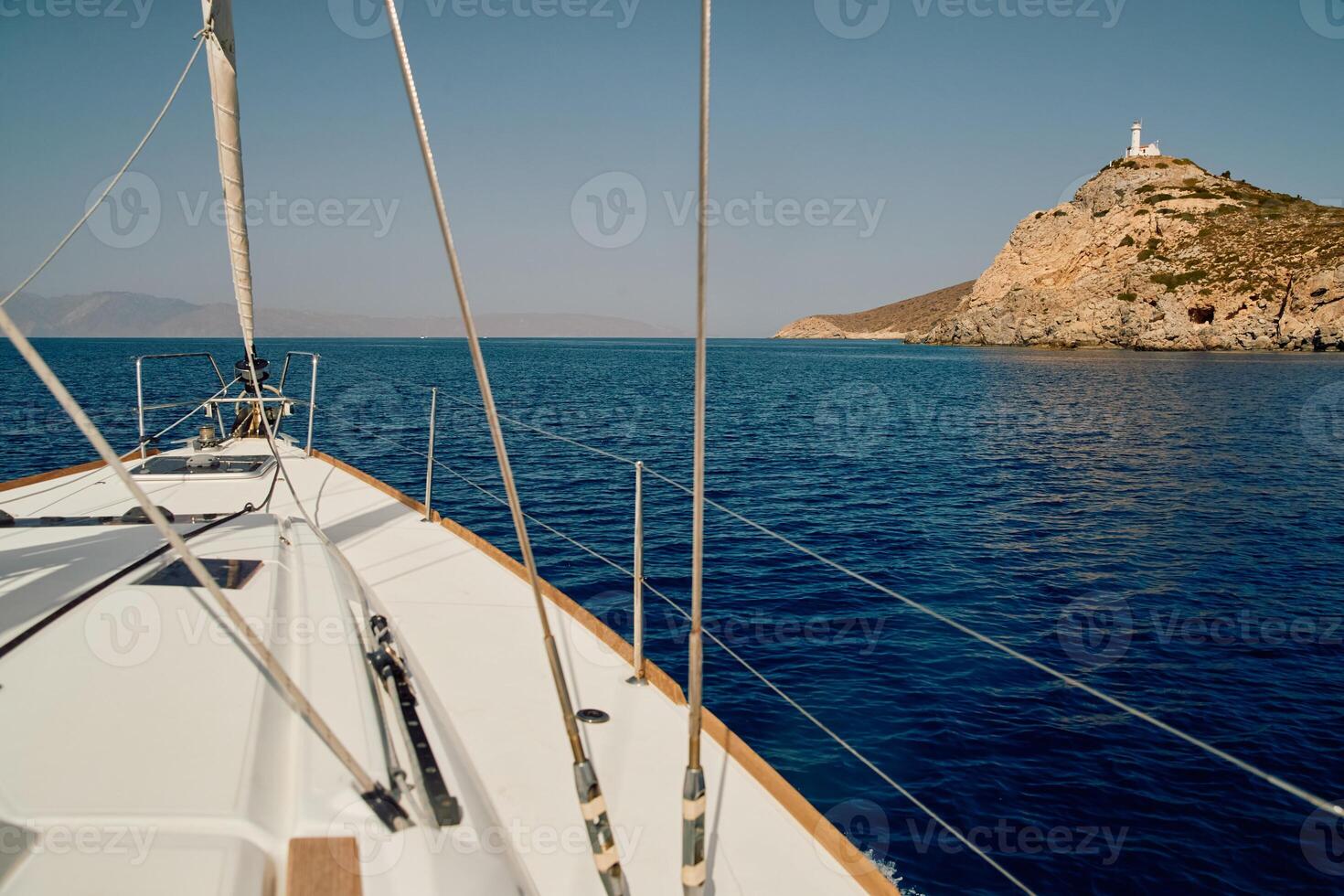 view from a sailing boat to an island with a lighthouse in clear sunny weather, the bow of the boat, the shrouds of the yacht, reflection on the water, mountains in the background photo