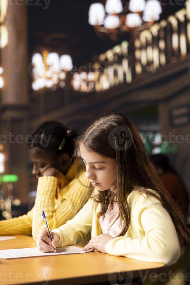 Preadolescent girls focused on literary analysis essay writing in the library, working together on school project and consulting the academic database. Academic child development. photo