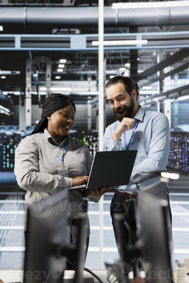 Cheerful team of engineers using laptop in server farm, analyzing data. Jolly teamworking employees examining infrastructure in data center using notebook, ensuring system integrity and security photo