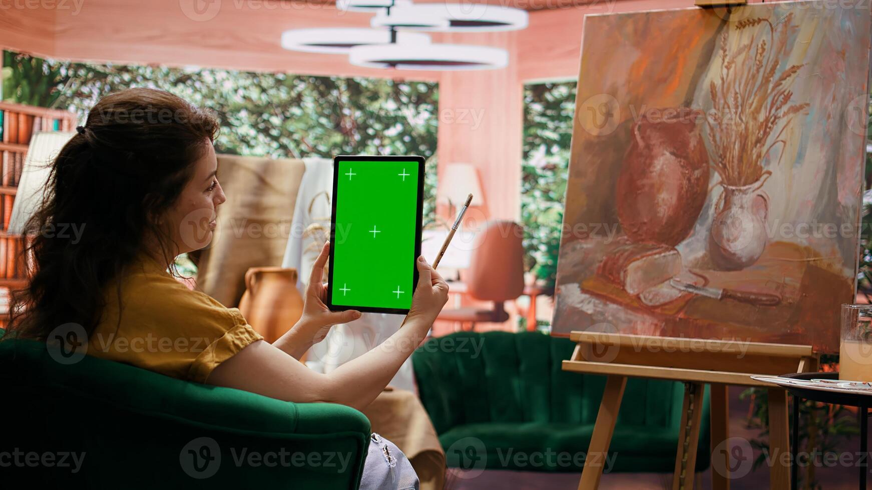 Artist using mockup copy space display in her painting workshop, looking at screen next to her rustic colorful masterpiece. Woman examining her finished artwork with watercolor paint. Camera B. photo