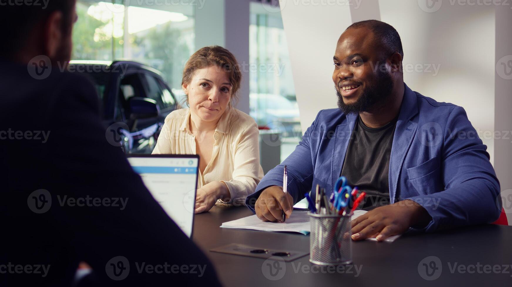 Customers in showroom completing credit application and financing paperwork after deciding to purchase new car. Salesman in dealership discussing financing options with clients, camera B photo