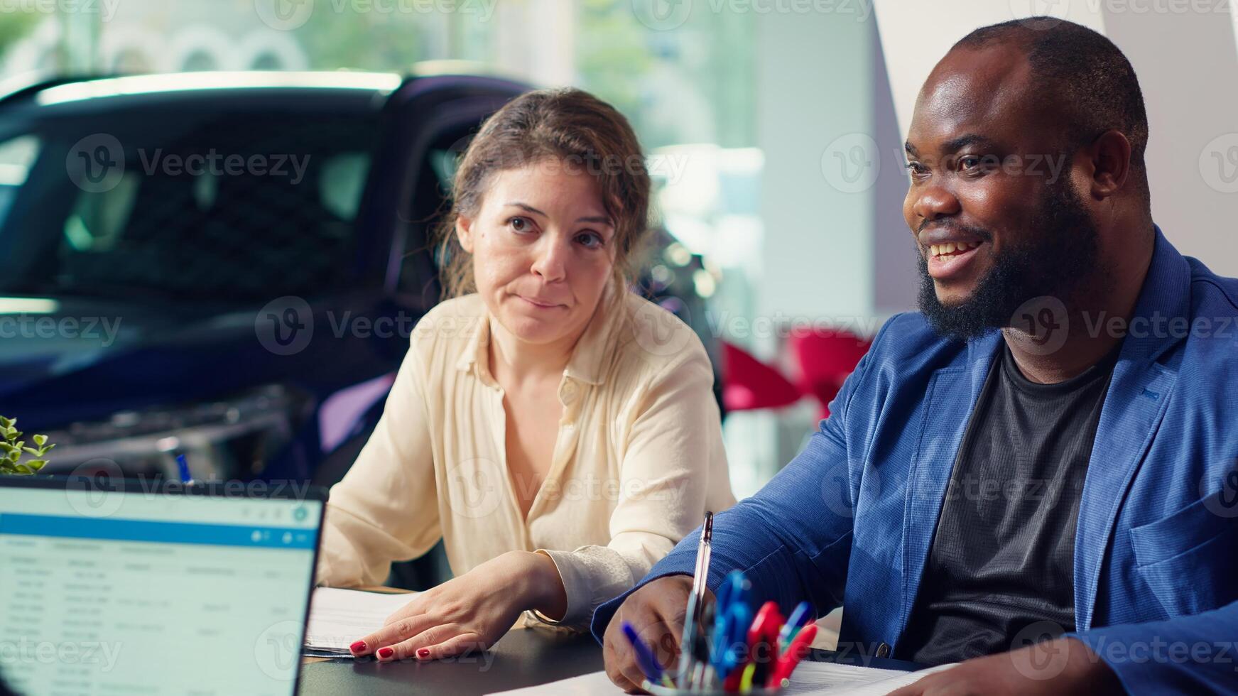 Customers in showroom completing credit application and financing paperwork after deciding to purchase new car. Salesman in dealership discussing financing options with clients, camera A photo