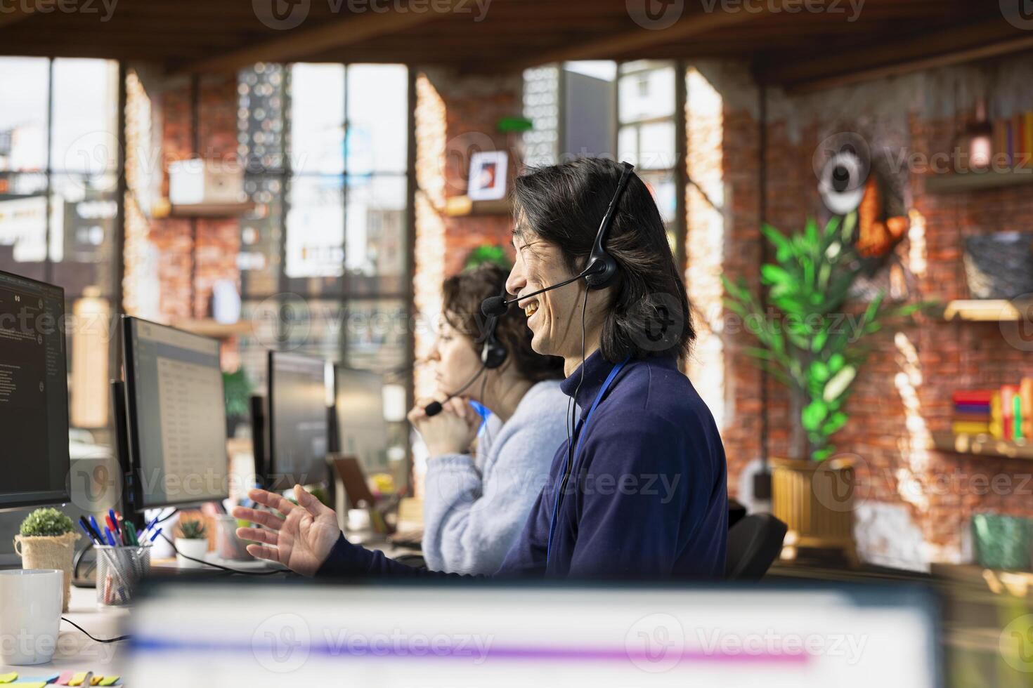 Call center agent laughing while talking with funny customer using headset mic while providing guidance. Customer center employee amused by comical client while providing technical solutions photo