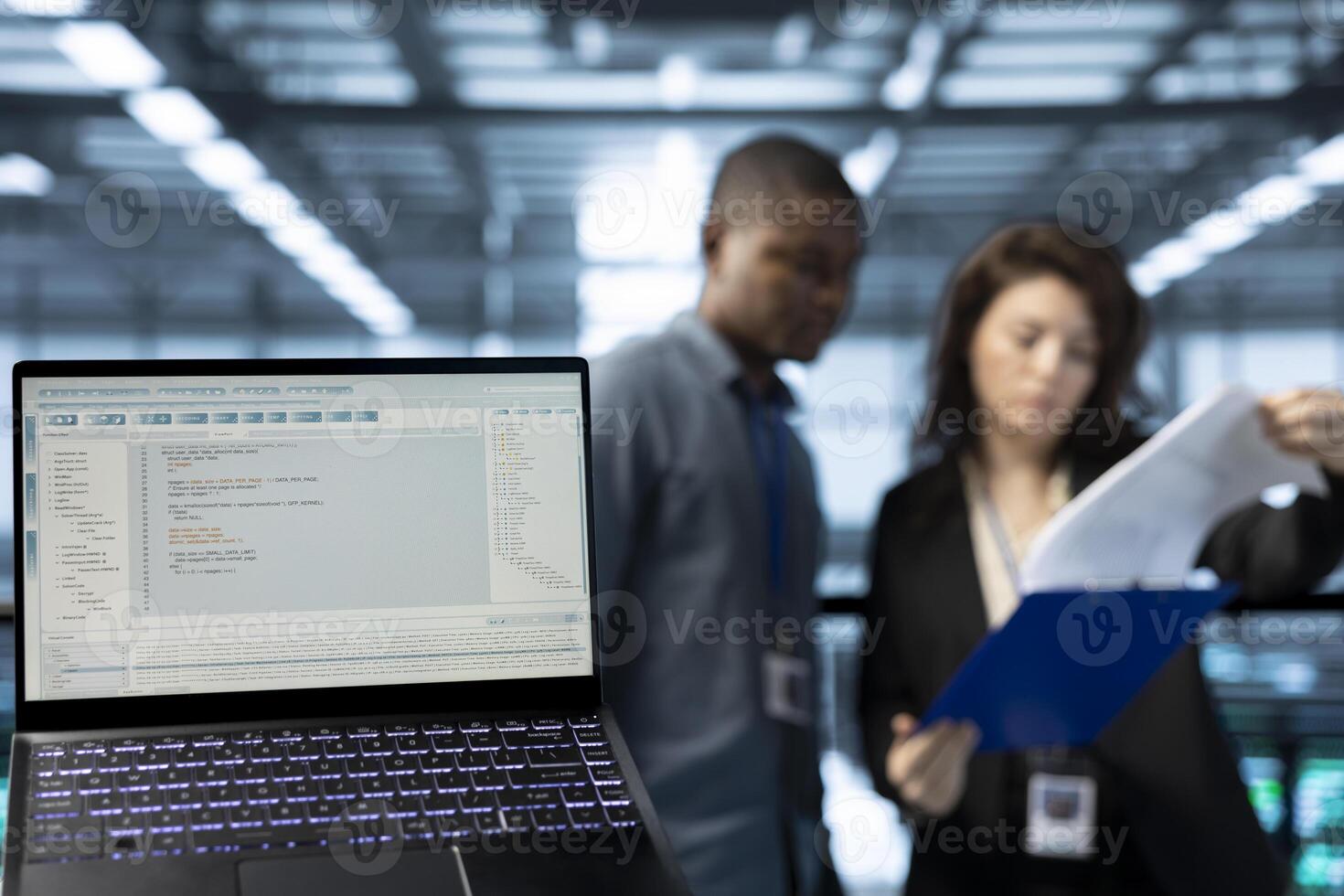 Close up of code lines on laptop screen in front of data center employees reading paperwork. Focus on programming language on notebook display next to server hub workers reading technical files photo