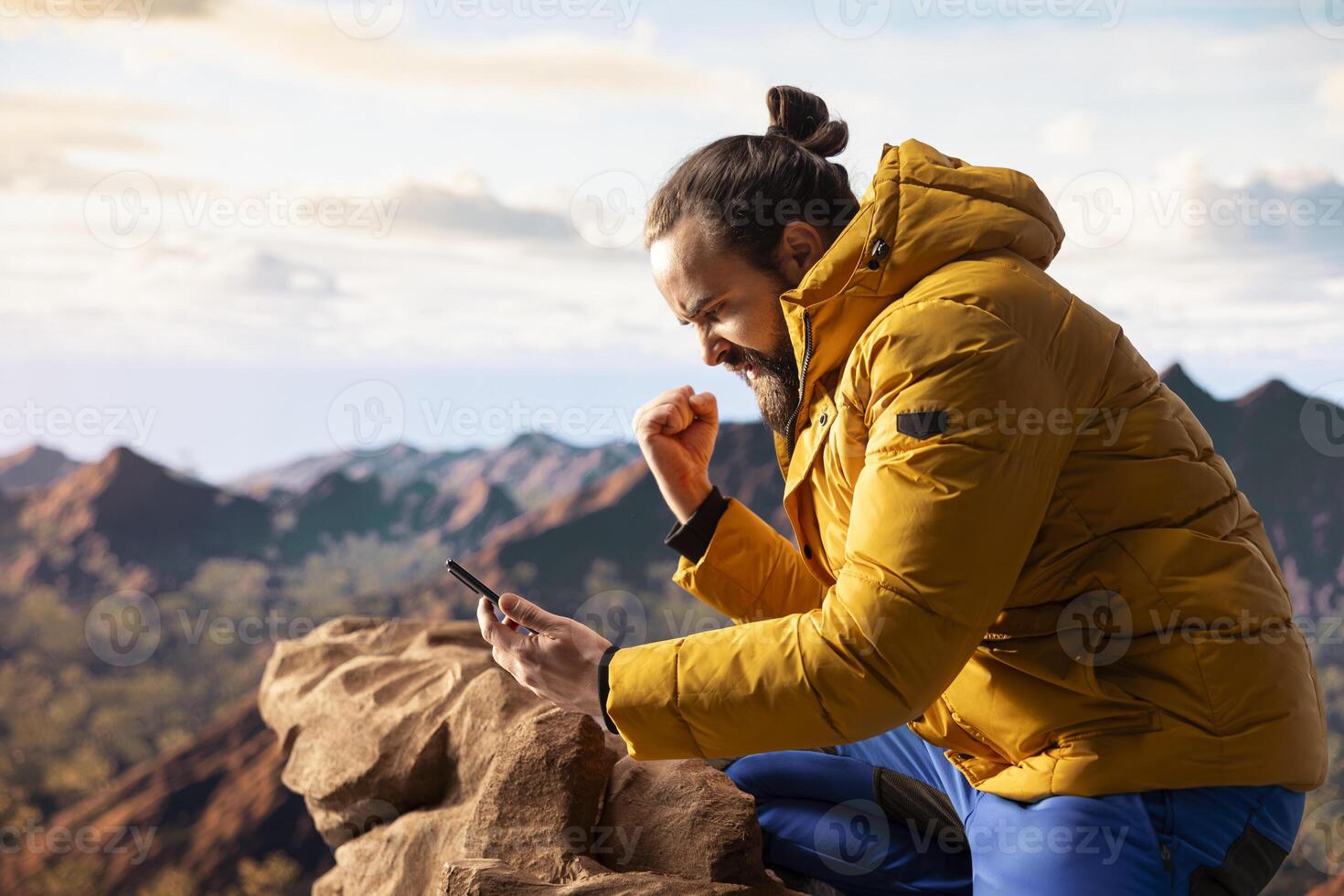Male hiker reading some good news and feeling joyful with his phone, being distracted by the strong signal and internet connection. Modern explorer cheering with excitement on mountain peak. photo