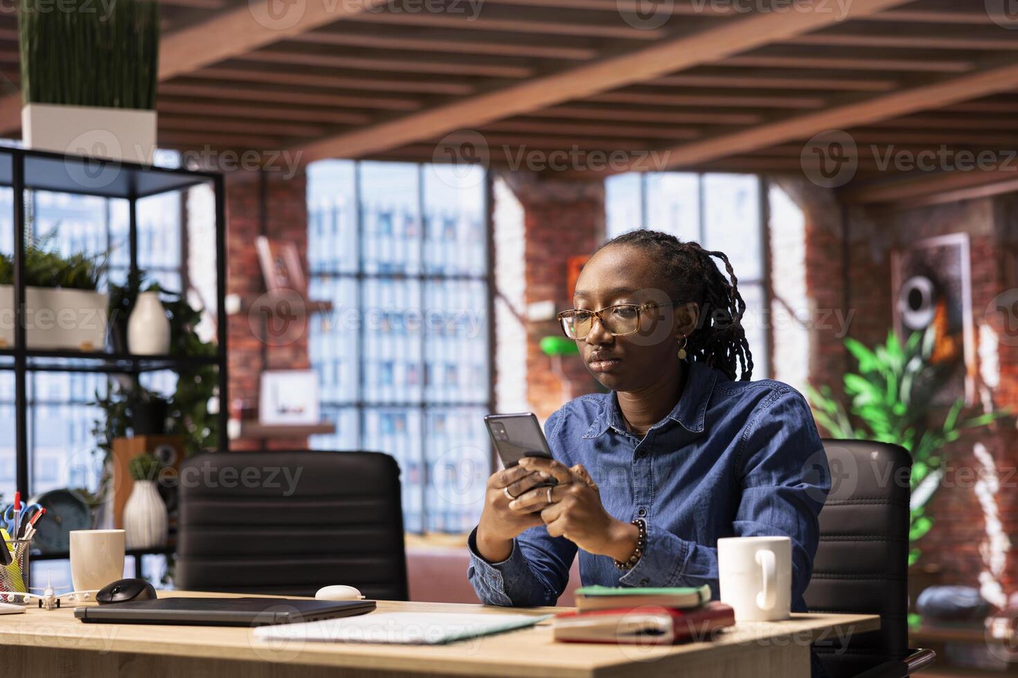 Female freelancer navigating on her smartphone at home office, browsing the web and demonstrating a lifestyle of productivity in a virtual workspace. African american woman multitasking. photo