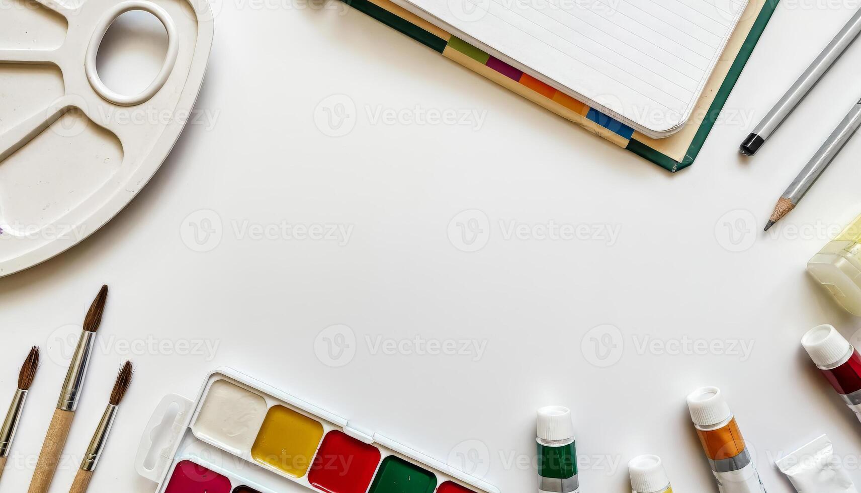 Creative workspace with watercolor paints, paintbrushes, color tubes, sketchbooks, and pencils arranged around a white background photo