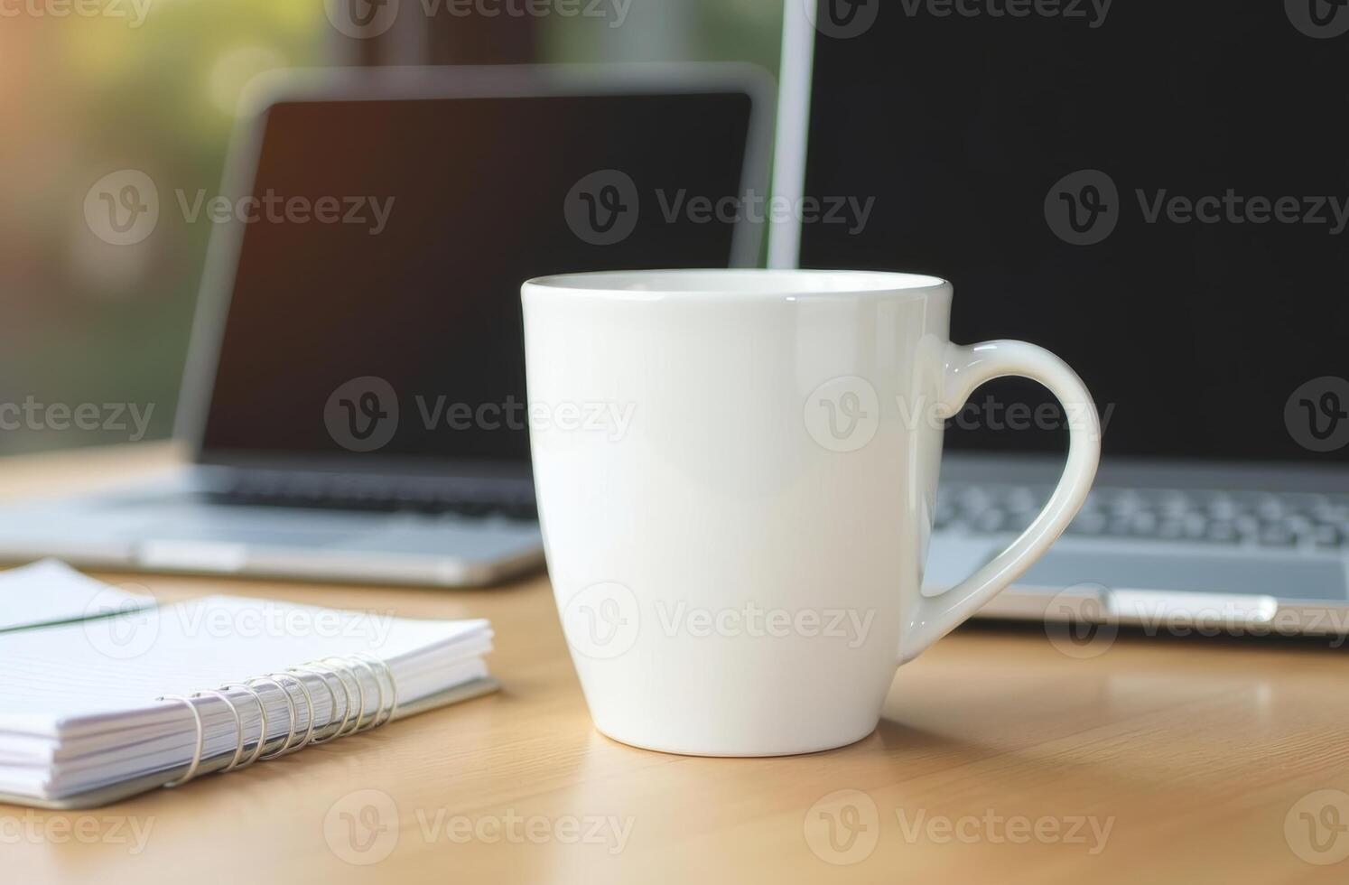 A white coffee mug on a wooden desk with a laptop, notebook, and a blank screen in a bright workspace during the day photo