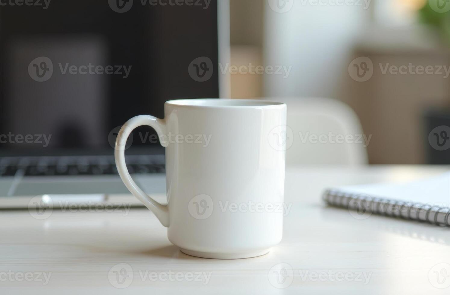 A plain white coffee mug sits beside a laptop and notepad on a wooden desk in a cozy workspace during daylight hours photo