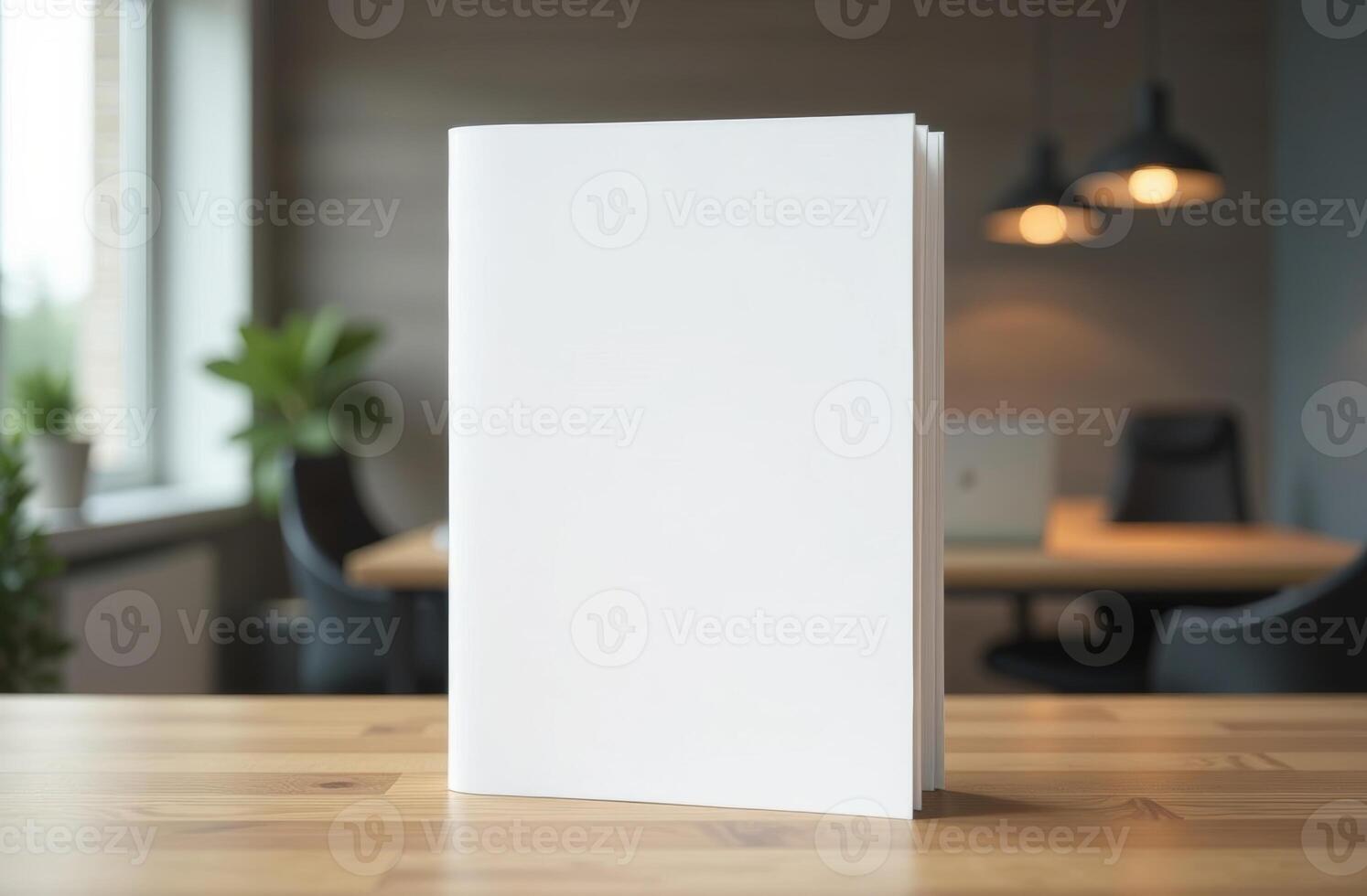 A blank notebook sits on a wooden table in a modern workspace with plants and warm lighting during the day photo