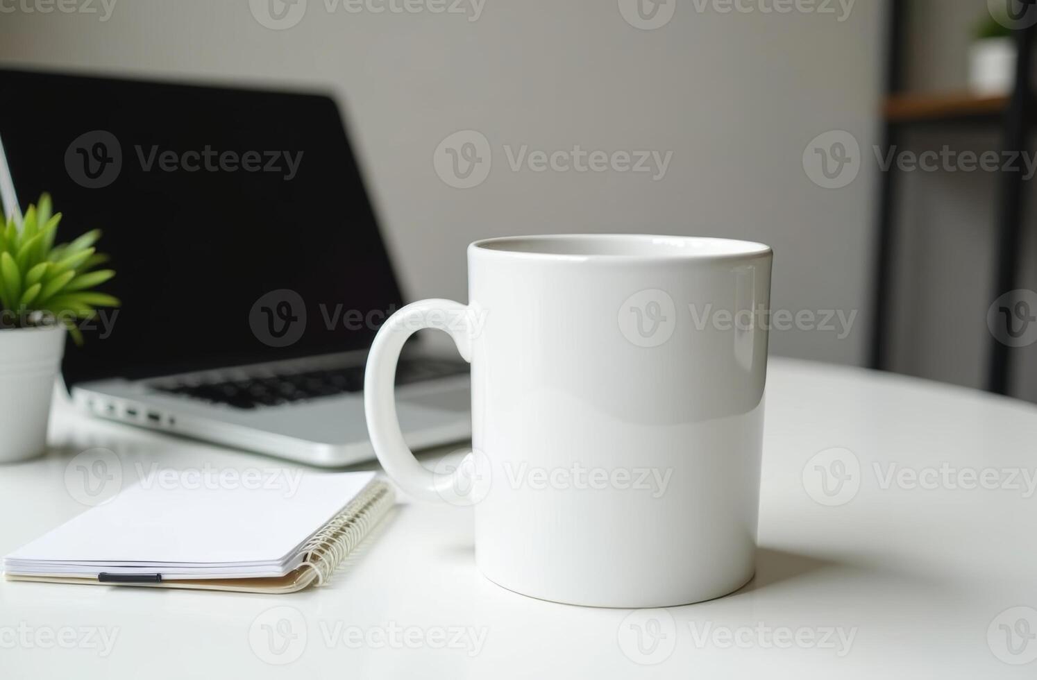 A white coffee mug sits on a table beside a laptop and notepad in a minimalistic workspace during daylight photo