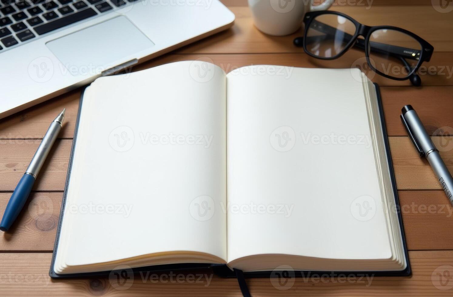 An open notebook, pens, and glasses arranged on a wooden desk next to a laptop and a coffee cup during a quiet work session photo