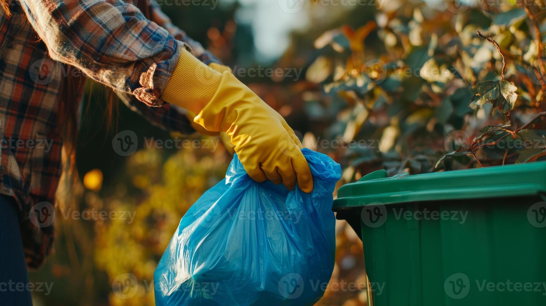Person wearing yellow gloves disposing of trash in green bin during daytime in a garden setting photo