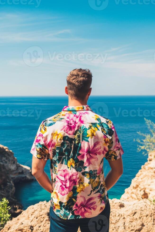 Man gazes at ocean view from rocky coastline, enjoying sunny day with bright floral shirt in paradise photo