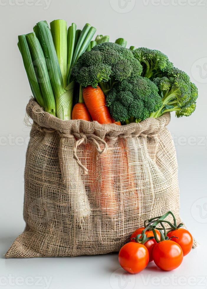 Veggies in a jute bag. A jute bag filled with fresh broccoli, carrots, and green onions sits next to vibrant red tomatoes on a neutral surface. photo