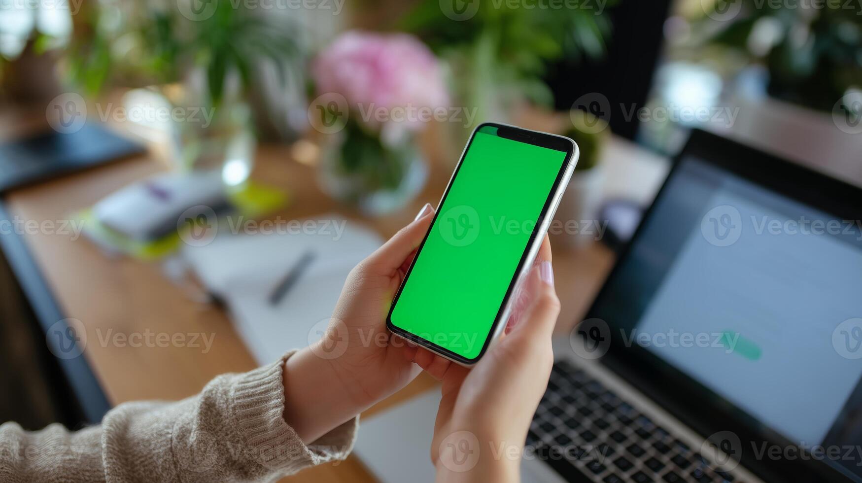 Mobile device held in hands with green screen, laptop and workspace in bright indoor setting during daytime photo
