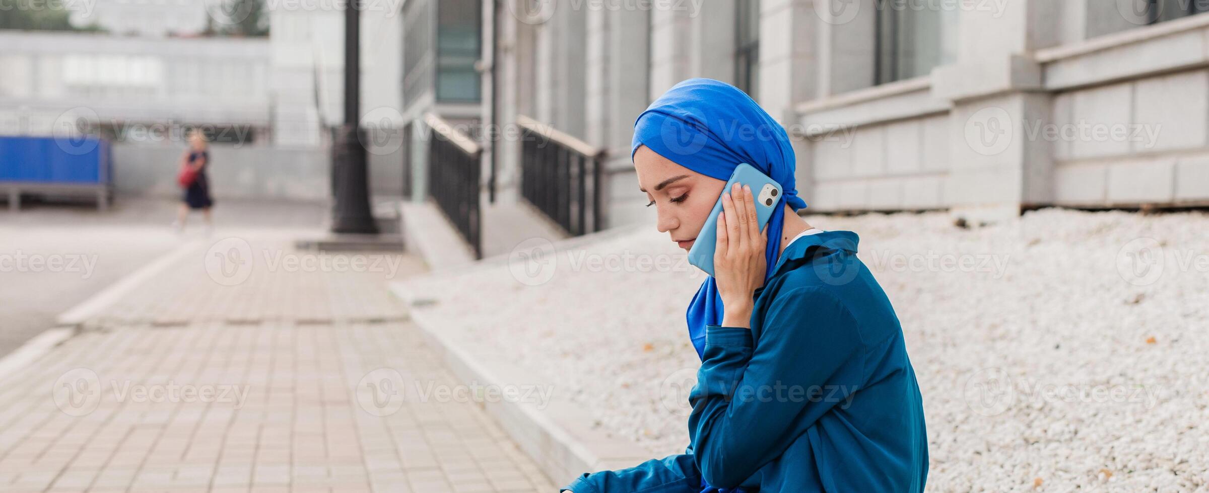 Young muslim woman in hijab speaking on the phone in urban surroundings. Communication, technology, and cultural diversity concepts. Banner with copy space photo
