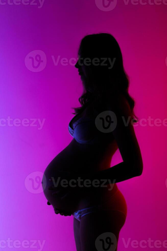 Beautiful young pregnant woman in lingerie is happily stroking her belly in studio in beautiful neon purple lighting. photo