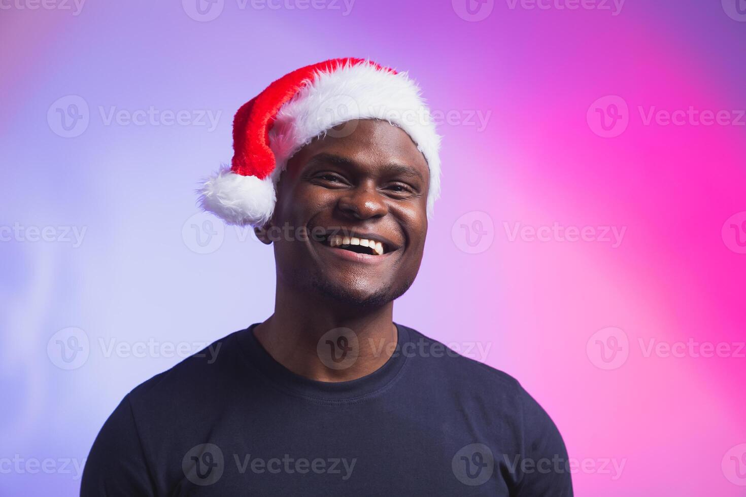 Portrait of positive smiling man in santa hat and casual t-shirt on colourful background, christmas time. Winter holidays and emotional people concept. photo