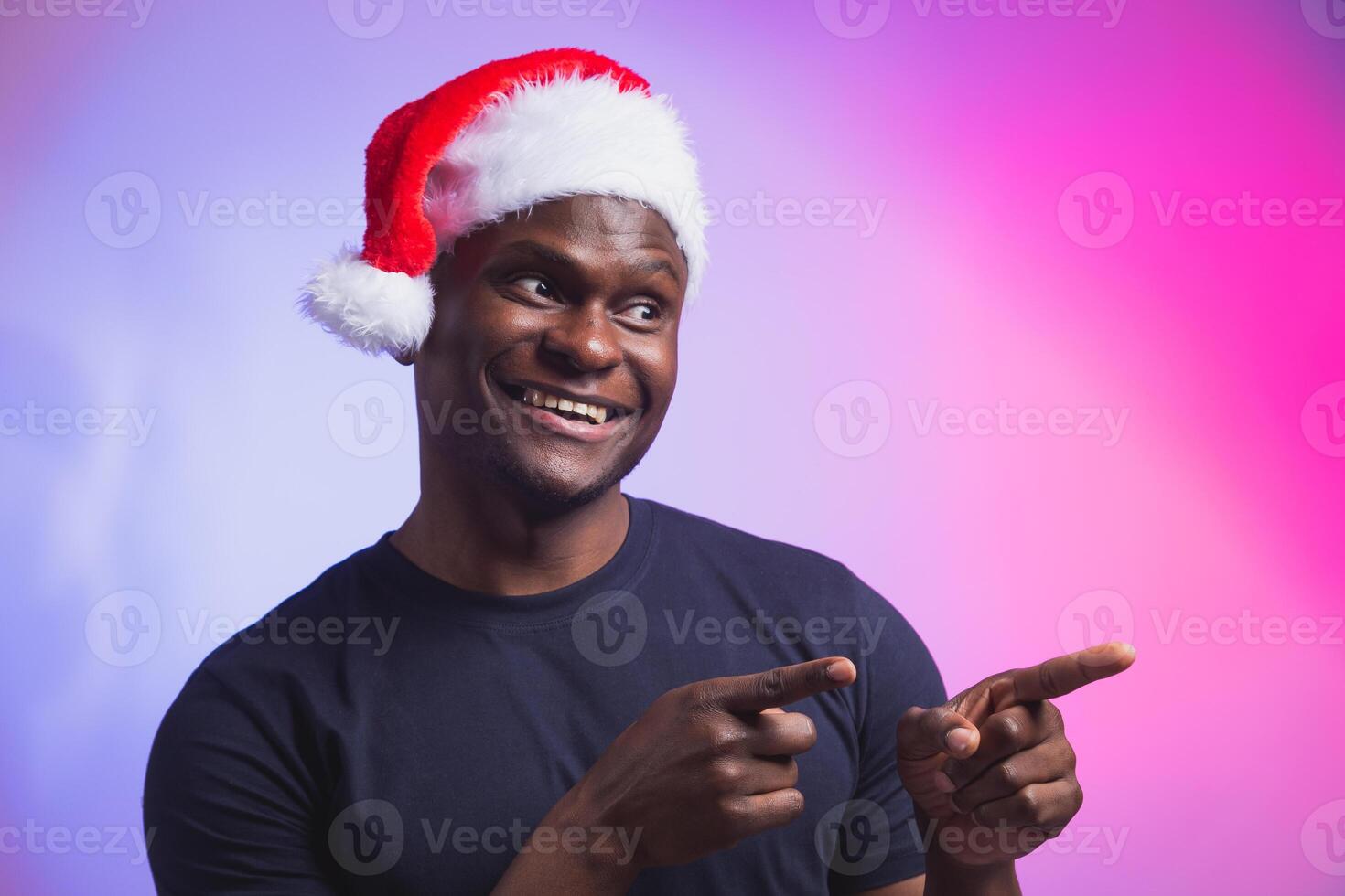 Portrait of positive african american smiling man in santa hat and casual t-shirt on colourful background, christmas time photo