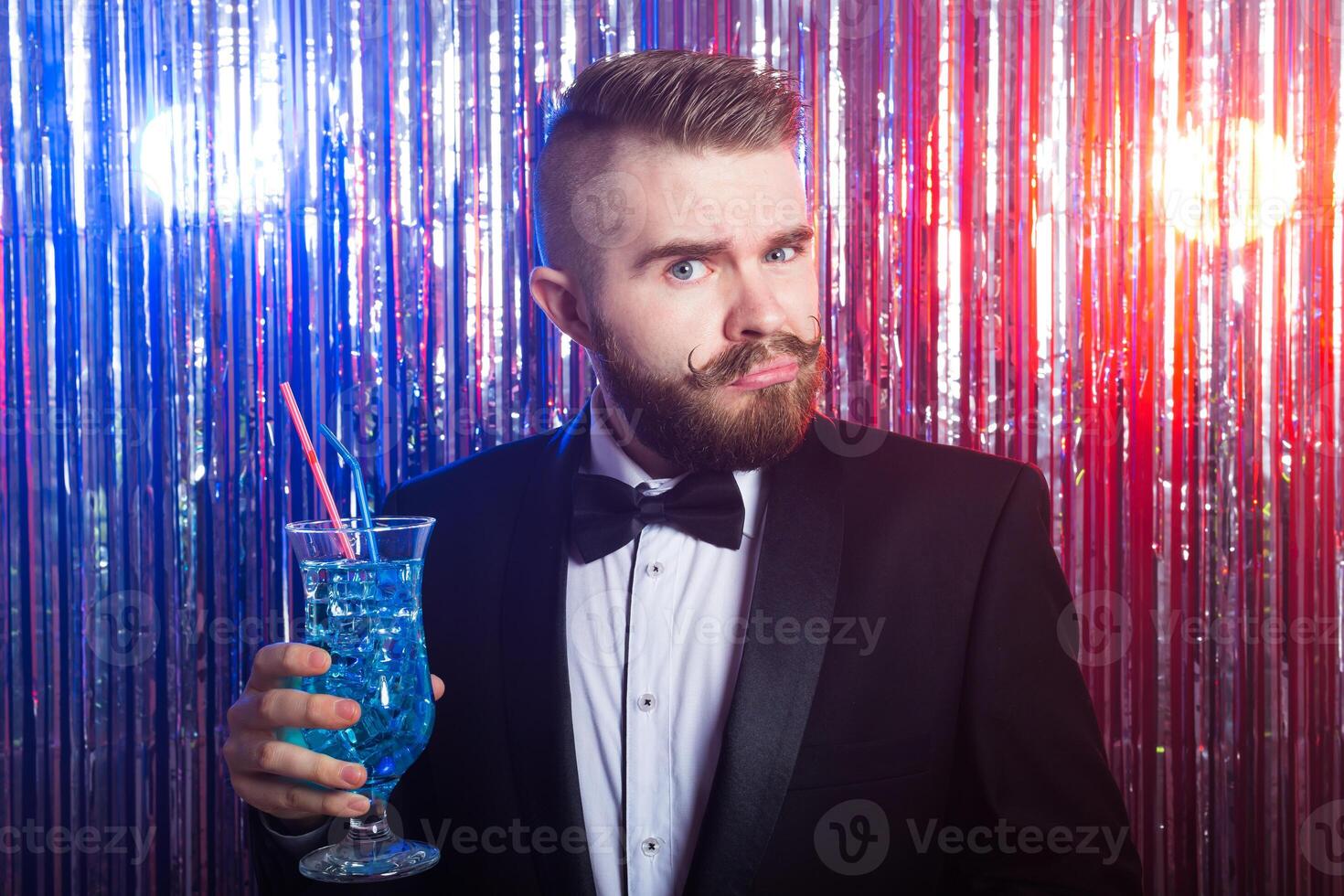 Club party, emotions and holidays concept - Portrait of elegant angry handsome man in a expensive suit holds blue cocktail on shiny background. photo