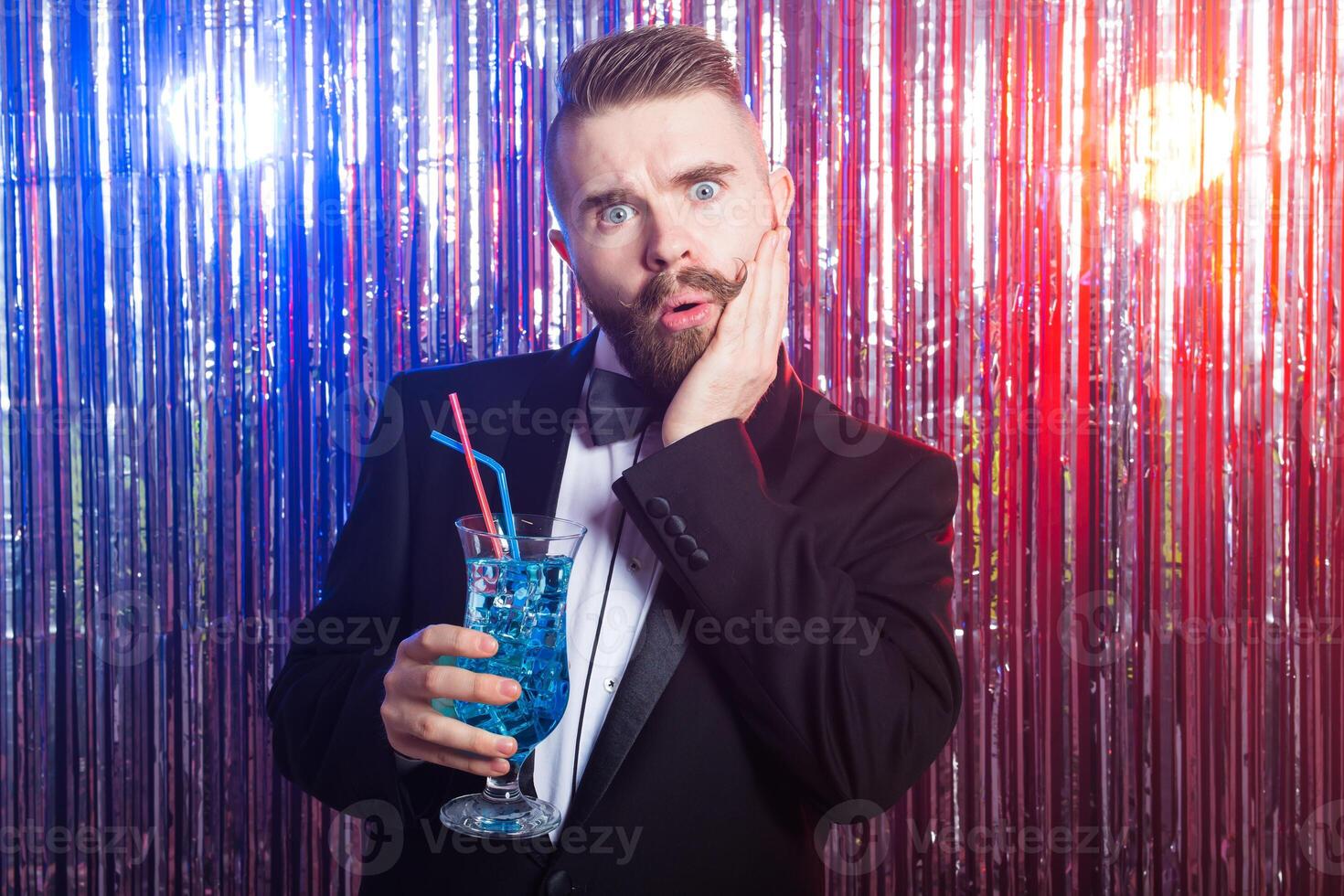 Club party, emotions and holidays concept - Portrait of elegant surprised handsome man in a expensive suit holds blue cocktail on shiny background. photo