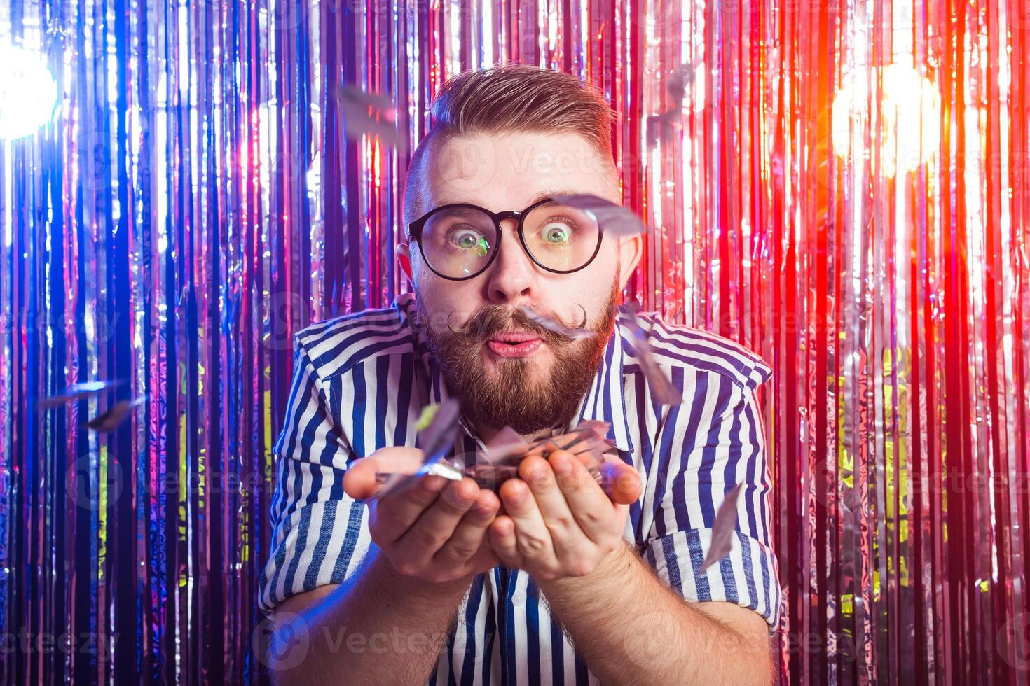 Party, carnival, holidays and fun concept - Fancy man blowing confetti against shiny background photo