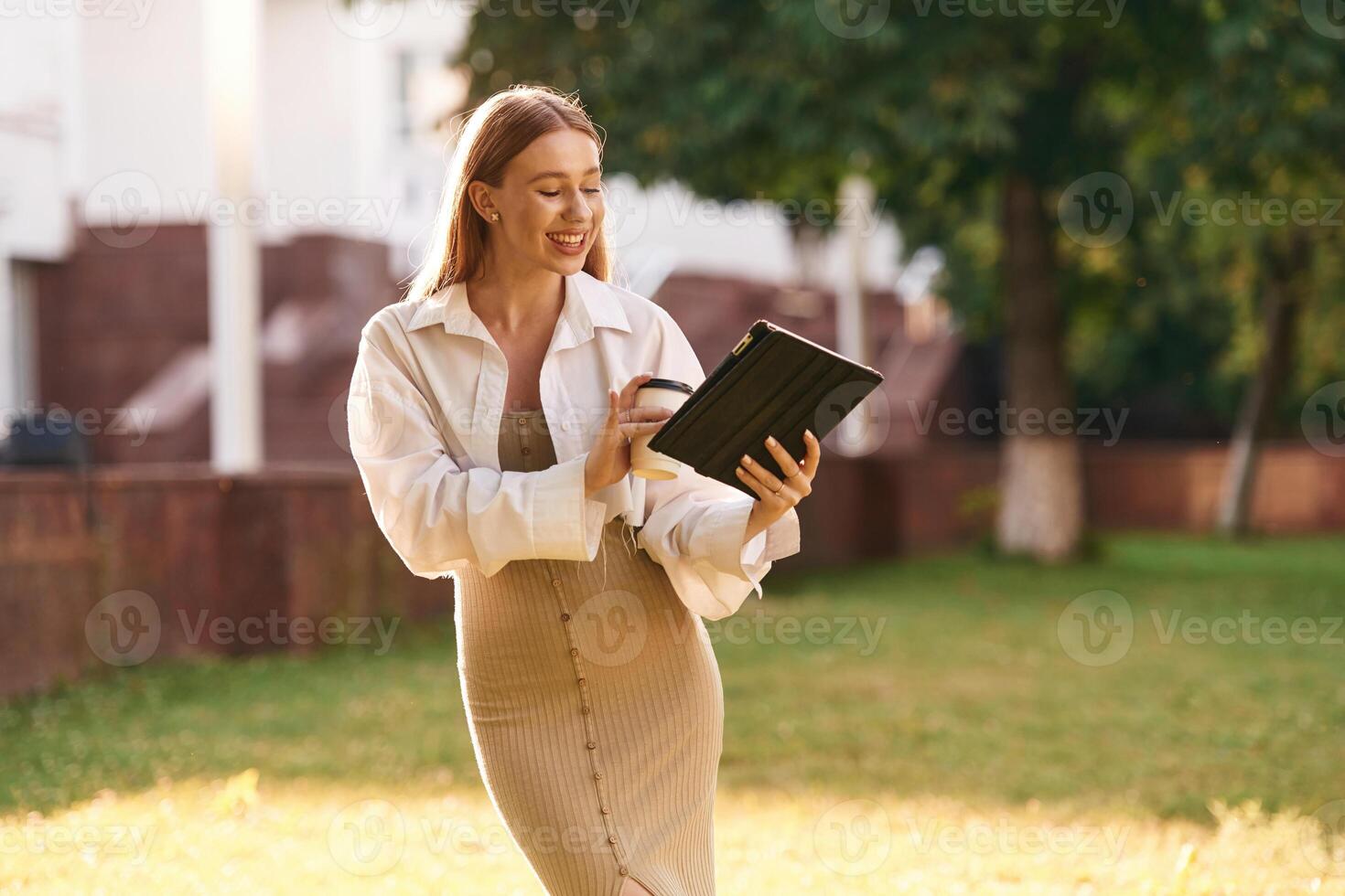 Digita,l tablet and cup of drink in hands. Beautiful female student is outdoors near the university photo