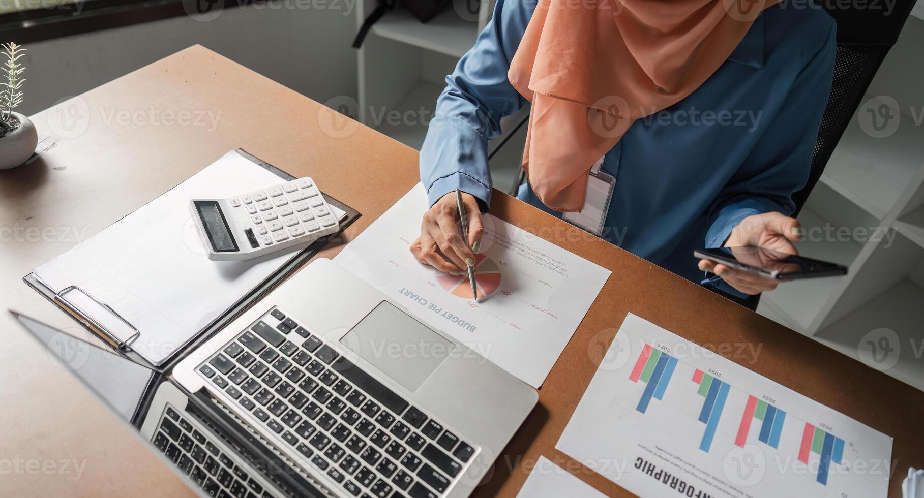 Muslim woman analyzing financial charts and using smartphone at her desk. photo