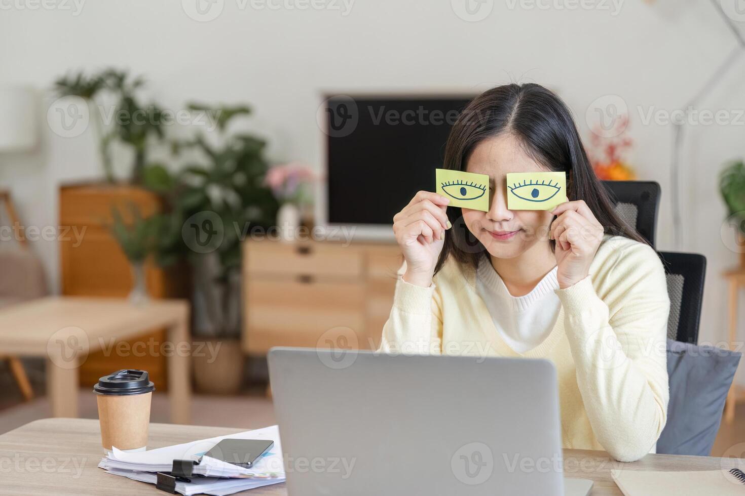 Playful Asian woman holding paper eyes while working on a laptop, showcasing creativity and fun in the home office. photo