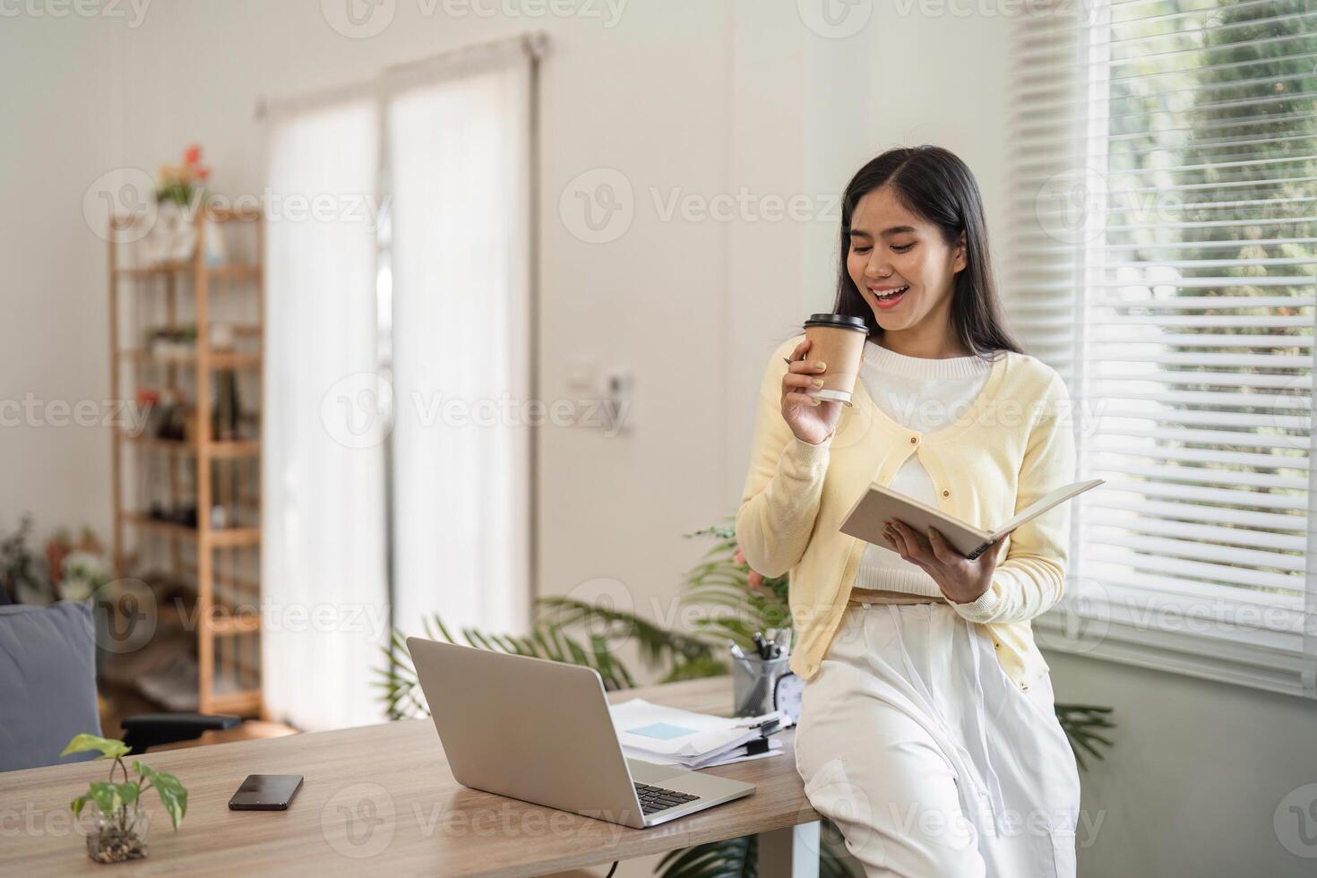 Young woman working from home, enjoying coffee while reading a notebook and smiling. photo