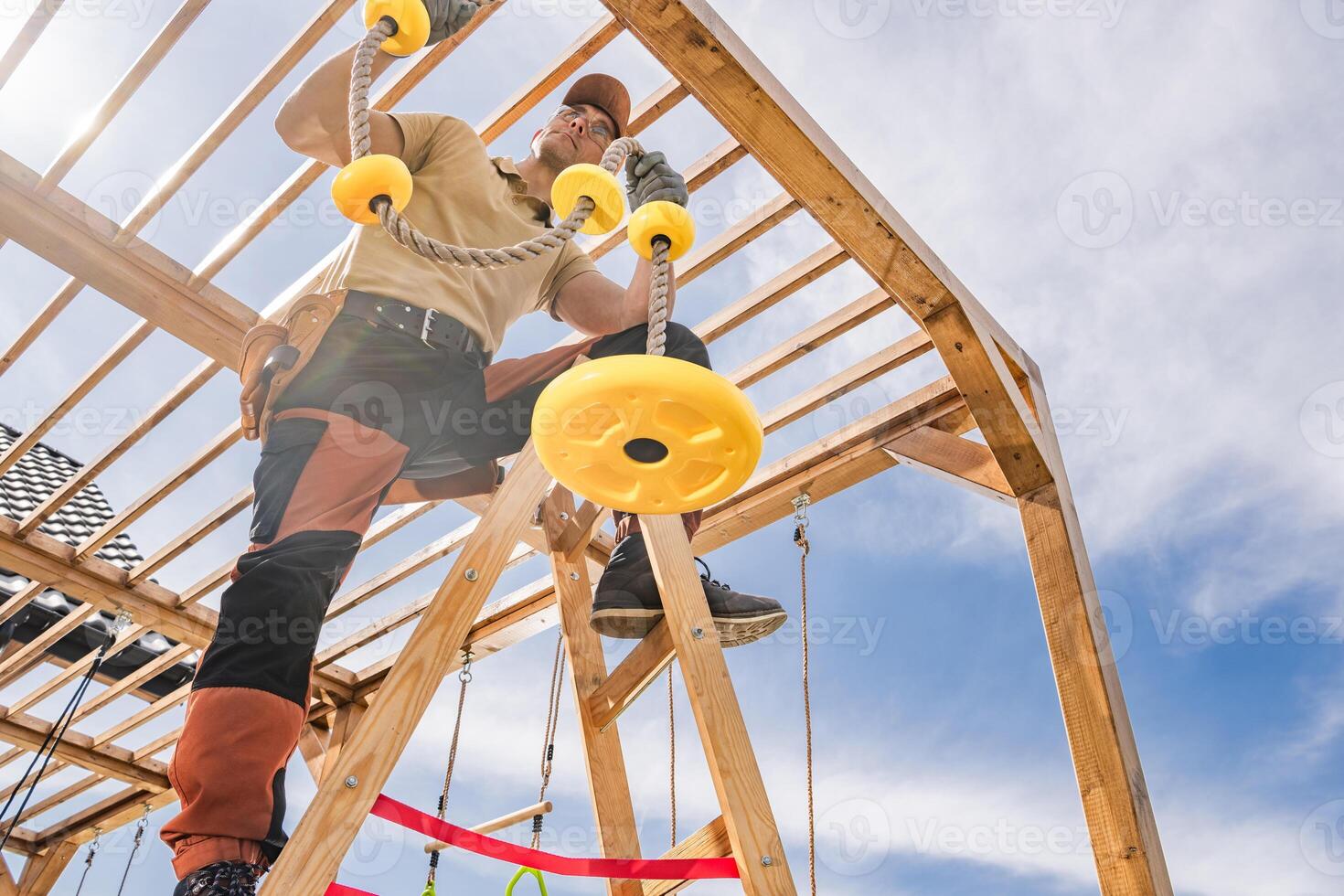Construction of a Wooden Playground Structure With a Man Installing Yellow Rope Grips in Bright Daylight photo