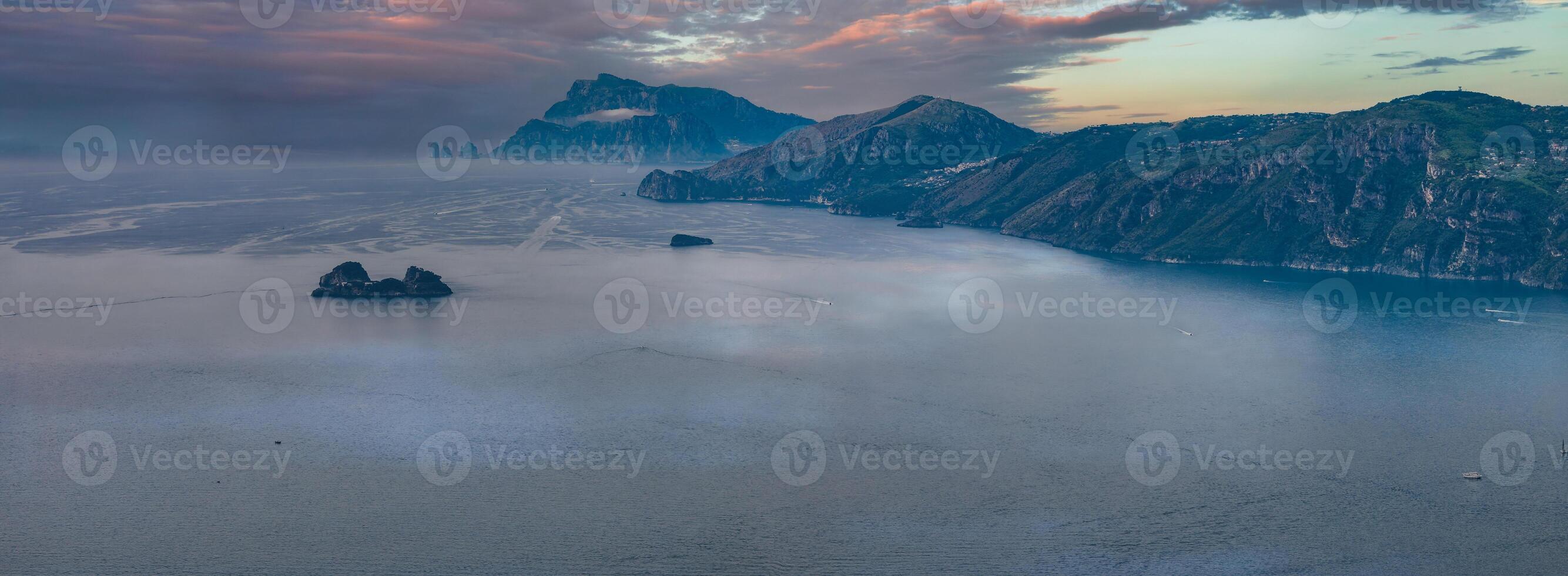 Aerial view of the Amalfi Coast with cliffs, rocky islets, and Capri's Faraglioni. The scene features a colorful sky transitioning at twilight. photo