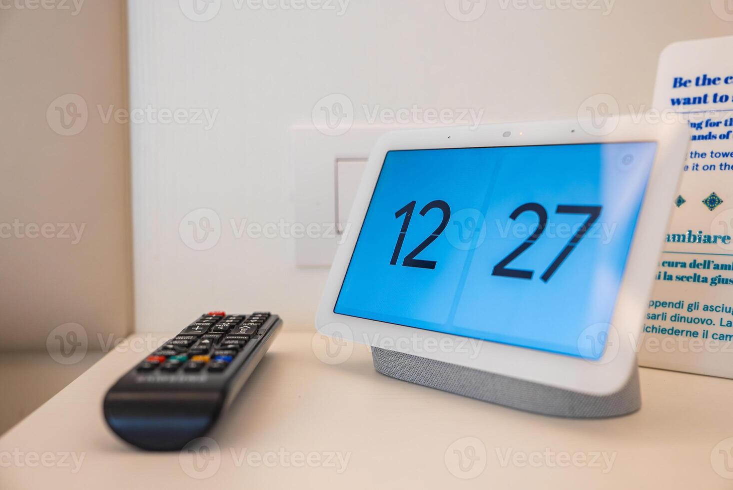 Modern digital clock displaying 12 27 placed on a white surface next to a black remote control. Background includes a printed sign and wall socket. photo