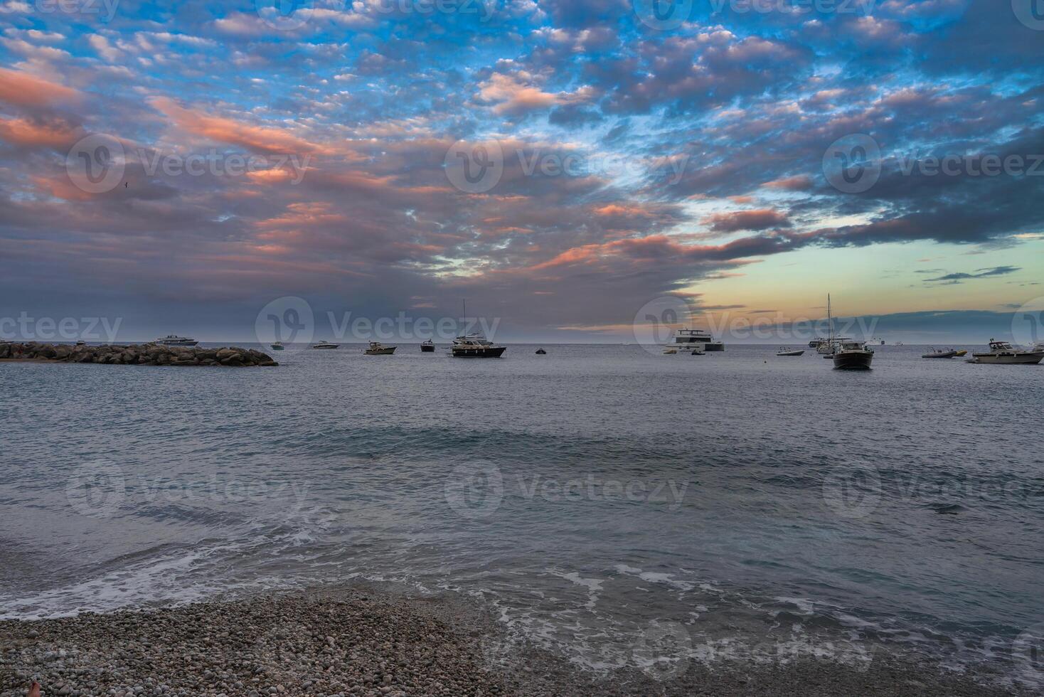 A serene coastal scene on Capri, Italy, with a pebble beach, calm waters, anchored boats, and a vibrant sunset sky with blue, orange, and pink hues. photo