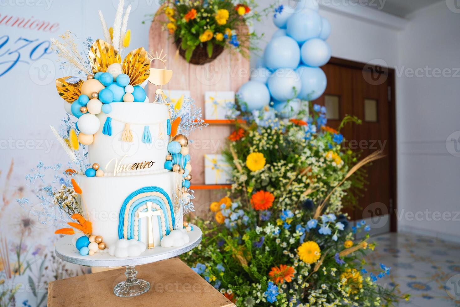 A multi tiered cake with blue, orange, and gold decorations, surrounded by floral arrangements and balloons, set in an indoor festive setting. photo
