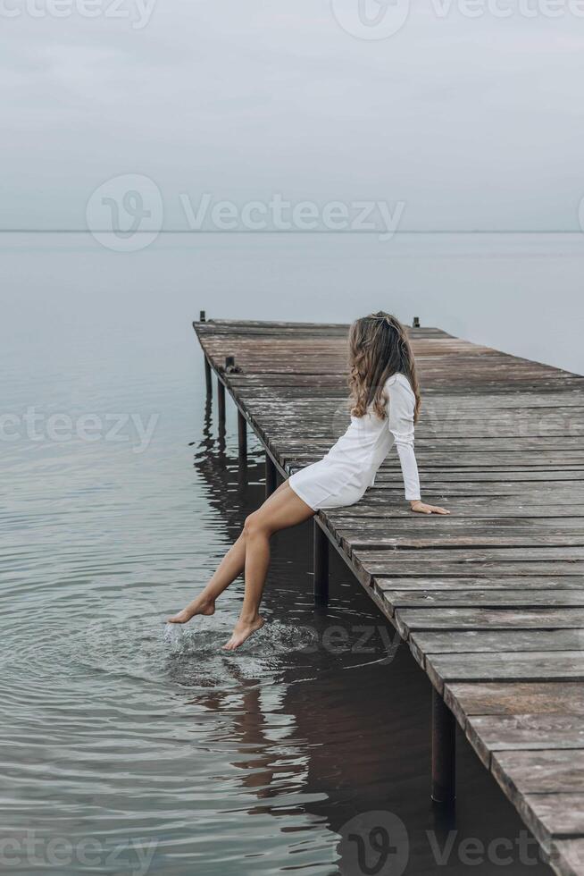 unrecognizable woman in a white dress, sitting on the edge of a wooden pier, dipping her feet into calm water with a light splash, gazing at the horizon under a soft cloudy sky, relaxation, travel photo
