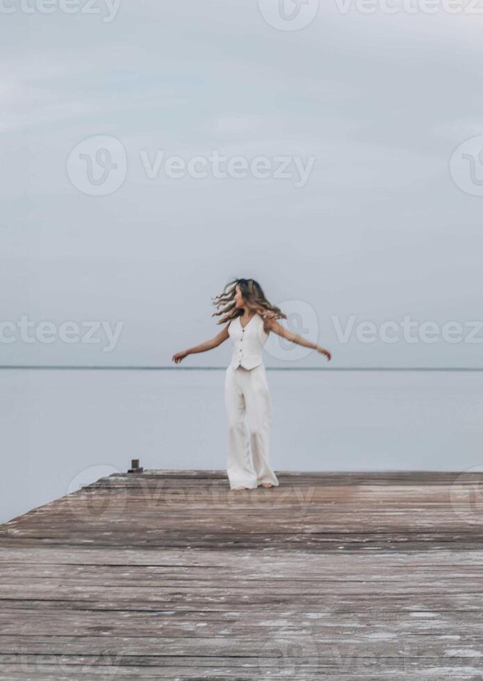 woman dancing barefoot on a wooden pier, long hair flowing as she twirls freely under a pale sky, surrounded by calm water, joyful expression in motion, freedom, wellness, motion blur photo