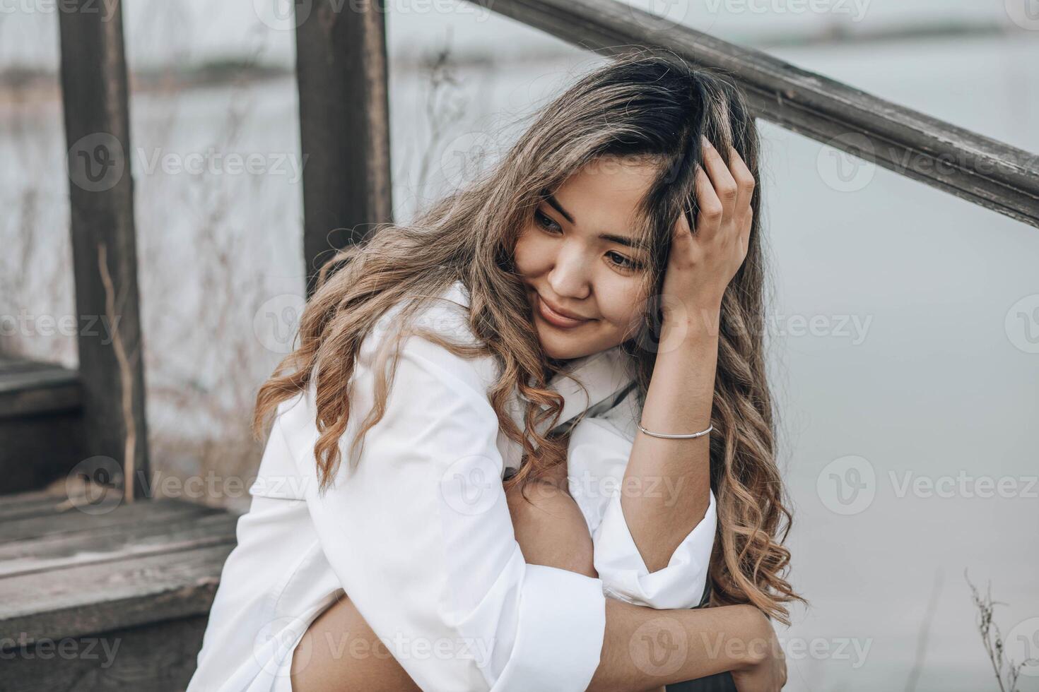Asian young woman with wavy hair on wooden stairs in white shirt, with hand in hair, peaceful emotional moment outdoors near water, mindfulness, feminine wellness, emotional balance photo