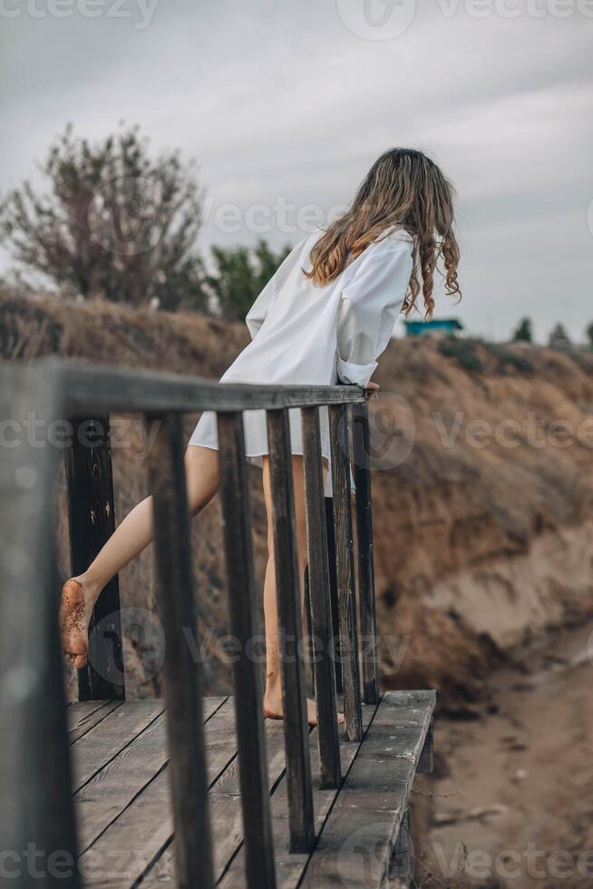 unrecognizable woman in white shirt standing barefoot on wooden platform with one foot raised, natural cliff landscape, back view, solitude, personal reflection, nature retreat photo