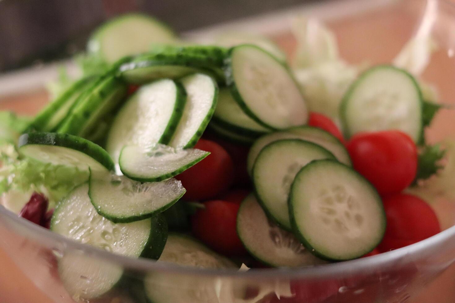 Fresh salad preparation with cucumbers, cherry tomatoes, and greens in a clear bowl on a kitchen countertop photo