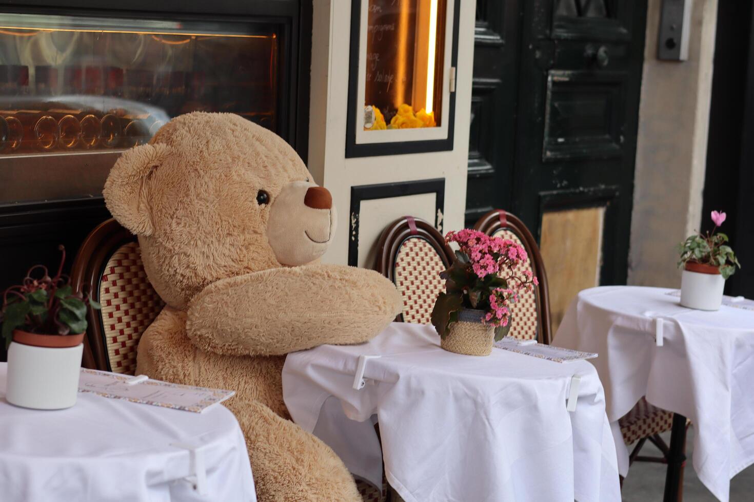 Large stuffed teddy bear sitting at a cafe table in a quaint outdoor dining area with flowers nearby photo