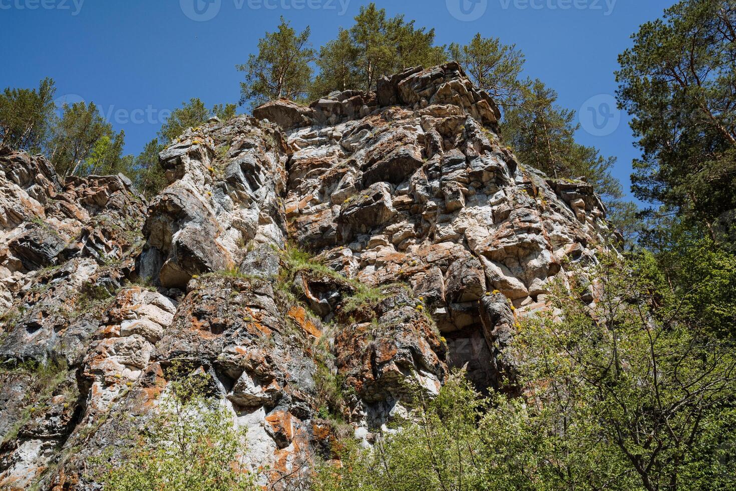A majestic rocky cliff adorned with vibrant trees scattered across its surface, complemented by a breathtaking blue sky gleaming in the background, creating a picturesque natural scene photo