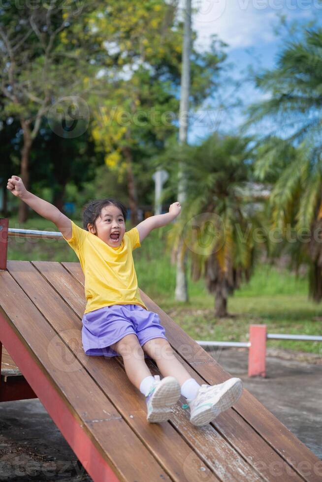 Joyful Asian Child Sliding Down Playground Slide on Sunny Day in Outdoor Park with Green Trees and Blue Sky in Background photo